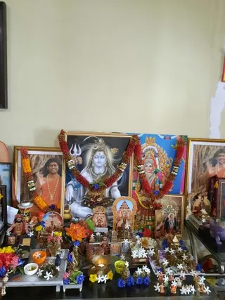 A home shrine on a glass-topped table against a pale yellow wall, decorated for daily puja. Central framed print shows a seated blue-skinned Shiva with trident and crescent moon, garlanded with red and orange flower strands. To the right is a framed image of a goddess in a golden crown with a blue backdrop, also garlanded. Framed photographs on the left and right show a smiling man in orange robes with a beaded necklace, one with a raised hand, set against a tree-trunk background; the left one wears an orange and yellow garland. Smaller gold-framed icons, a golden temple-arch picture, a brass lamp, small bowls of offerings, white jasmine flowers and blue and yellow blossoms surround the altar. No people are physically present.
