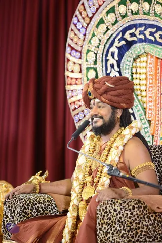 Swamiji (probable) seated on a leopard-print chair in front of a maroon curtain and an ornate, jewelled golden-green-blue throne backdrop. He wears a rust-brown turban with a blue gemstone brooch, a rust-brown dhoti-style wrap, multiple long gold and white flower garlands, gold armlets and bangles, and a gold earring. He has a full beard and long hair, smiles while speaking into a black microphone on a stand, and rests his hands on the chair arms. A gilded lion-like statue is partly visible at the left edge.