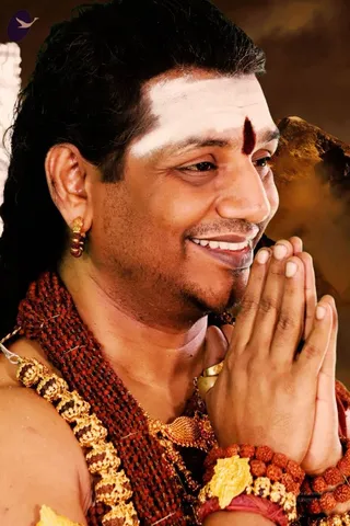 Close-up photograph of a smiling man with short dark hair, a white horizontal forehead mark and a red bindi-style tilak, his palms pressed together in anjali namaskar. He wears a thick rudraksha mala across his chest, a gold-and-bead ornamental necklace, a small gold earring and rudraksha bracelets on his wrists. A brown, mountain-like background is visible behind him.