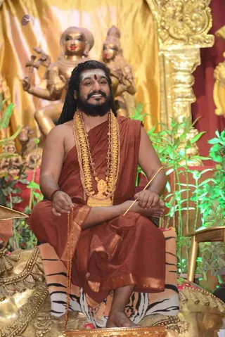 A photograph of a smiling bearded man with long dark hair seated on a golden throne draped with a striped tiger skin. He wears a rust-orange shawl draped over bare shoulders, a heavy gold necklace with a large gold pendant, and rudraksha beads. He has a white and red tilak on his forehead, holds a mala in one hand and a slender golden staff in the other. Behind him stand golden deity statues against a gold draped backdrop, with green potted plants flanking the throne.