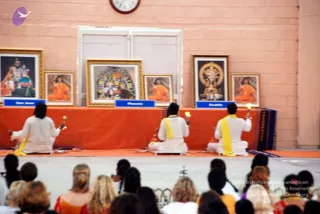 A photograph of a ritual ceremony in a hall with pinkish walls and a wall clock. Three people in white garments with yellow sashes sit on an orange-draped stage, each holding a lit oil lamp aloft. Behind them stand several framed portraits and paintings on an easel-like display, including deity images and a framed image of a seated figure in saffron robes. Blue name placards on the stage edge read San Jose, Phoenix and Seattle. A seated audience of many people in the foreground, mostly from behind, fills the lower half of the image, with women in orange and white visible.