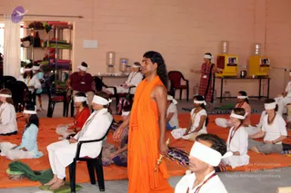 A photograph of a meditation session in a plain hall with pale pink walls and water jugs along the back. Many participants sit cross-legged on orange mats, most wearing white headbands over their eyes and white or light clothing with beads. A man in a long saffron-orange robe with a swept-back long hairstyle and a gold-handled staff stands at the centre, appearing to lead the session. Another person in a red and white checked wrap stands at the right edge. Shelves with folded coloured cloths sit at the left, and a yellow dispenser stands on a table at the right. The appearance-based label for the standing man is Swamiji (probable): he has long swept-back hair, saffron robes and a gold-handled staff, though the features are not clear enough to confirm.
