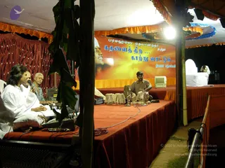 A night-time musical performance under an orange-trimmed canopy on a stage draped in maroon cloth. On the left, a singer in white clothes holds a microphone and sits at a harmonium, with a man in a grey shirt beside him. In the centre, a musician in a dark shirt plays a pair of tabla drums on the floor. A large orange banner behind the stage shows a smiling man's face and Tamil and English-style lettering. Banana-leaf decorations, a cooler, and a white covered seat sit at the right edge.