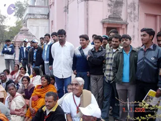 A photograph of a crowd outside a pink sandstone temple building with a small white and pink shrine structure at left. A standing row of young men in casual jackets, sweaters and jeans fills the right side, several smiling. In the foreground, seated people include an older woman in an orange sari, a man in a white shirt with a red lanyard and a shawl, and a woman in a white sari with red border. Some people wear white caps and red tilak-style marks.