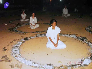 Night photograph of seated practitioners meditating on sandy ground inside concentric circles outlined with ash or burnt material. In the foreground a young woman in white clothes and a white shawl sits cross-legged with hands folded and eyes closed. Behind her, a woman in white with a beaded necklace sits in a similar circle, and further back several more people in white sit in meditation within the circles. Dim lights are visible in the background.
