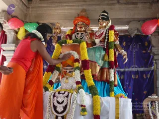 A photograph of a festival scene with two large golden utsava murti figures seated on a platform, one wearing an orange turban and the other a dark headdress with a teal and gold sari. Both are draped with yellow and red marigold garlands and layered white and gold necklaces. In the foreground a person in an orange sleeveless top and orange cloth leans over the platform, reaching toward the decorated base where a white cloth, a silver-and-red jewelled breastplate and marigold garlands are arranged. Behind them hangs a purple cloth with gold embroidery including a trident motif, and colourful balloons float at the upper left and right. A partially visible person stands at the left edge.