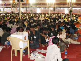 A large indoor hall with a red carpet packed with seated attendees, mostly men and young men in white shirts, dhotis and dark shirts, sitting cross-legged in dense rows. A white cardboard box stands on a beige plastic chair in the foreground beside scattered papers and water bottles. Overhead are bright lights, hanging decorative tassels and banners, and speakers on poles. Many attendees hold papers and write or read. No speaker or central figure is clearly visible in this frame.