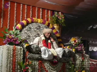 A man seated on an ornate zebra-striped throne decorated with long garlands of yellow, purple and red flowers and white jasmine strands. He wears a cream-coloured turban, a white-and-cream patterned kurta and white dhoti, with a red and white flower garland around his neck, and he smiles while raising one hand near his face. The throne sits on a flower-covered dais in front of an orange draped canopy with hanging white flower strings, potted pink and purple floral arrangements and a golden vase nearby. A stage setting for a festival day.