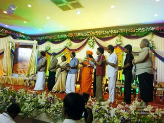 A stage decorated with flower garlands in white, orange and purple, draped gold and purple fabric, and multicoloured lighting. A large book cover with orange and gold design sits on an easel at left, reading a Tamil title. Eight people stand in a row facing right, toward the book. A man in a saffron robe with an orange head wrap stands centre, hands raised together. Others wear white dhotis and shirts, a woman wears a cream and gold sari, and a man on the right wears a light grey shirt. Photographers' silhouettes and white and purple lilies fill the foreground.