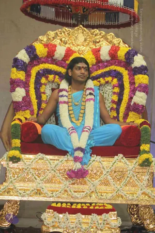 A photograph of a man seated cross-legged on an ornate gilded throne decorated with a flower-covered backrest of yellow, white, purple and pink blossoms. He wears a turquoise sleeveless garment with matching trousers, a heavy gold multi-bead necklace and a red bindi on his forehead. A long white and pink garland hangs over his shoulders and chest, and a flower bundle rests at his feet. Gold-framed floral footstool and a red-and-gold canopy with tassels frame the scene. Appearance-based candidate: Swamiji (probable), seated on the throne with long swept-back hair, gold ceremonial necklace and turquoise clothing.