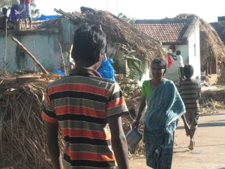 A photograph of a cyclone-damaged village street in Kuttiyankuppam, Pondicherry. A boy in a red, black and white striped polo shirt stands with his back to the camera, facing an older woman in a teal-blue striped sari and green blouse who smiles and carries a metal pot. Behind them, thatched roofs are torn and collapsed, with dried palm fronds and debris piled beside a whitewashed wall and a red-tiled house. A child in a striped shirt walks in the background, and a figure stands on a rooftop at the upper left.