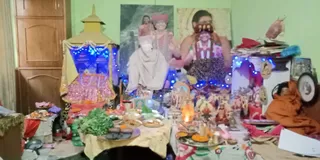 Indoor shrine table in a room with pale green walls and wooden cabinets, set up for a celebration. Two large printed portraits hang on the wall: one shows a man in a white shawl seated, another shows a smiling figure in red and white ritual clothing with a garland; a third portrait shows a woman with a decorated headpiece and multi-strand garland. Blue fairy lights frame the shrine. A small golden pagoda-style canopy stands at left. The table holds offering trays with green leaves, fruit, sweets and marigold petals, an oil lamp burning, small brass and clay idols, and a kalasha-like vessel. At right a seated figure in orange robes sits on the floor beside a stack of books. No people are clearly standing in the room apart from the seated figure, and the portraits are printed images.