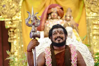 A photograph of a smiling bearded man seated on an ornate silver-white throne before a golden yellow backdrop. He wears a rust-red saffron-toned cloth over the shoulders, a rudraksha mala draped across his chest, and a pink-and-white flower garland. He holds a tall silver trident with a red stone in his right hand, and has white vibhuti horizontal markings with a kumkum dot on his forehead. Behind him, two deity murti figures in red and gold garments are visible, and green leaves frame the lower edge. a person cues: long swept hair, rudraksha mala, saffron-toned clothing and a ceremonial throne seat.