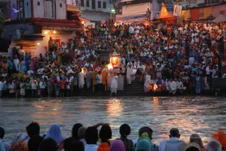 Evening Ganga Aarti at a riverside ghat with a large crowd densely packed on the stone steps and along the embankment. Several men in white dhotis and kurtas stand near the water's edge and on the steps, some holding lit oil lamps and a tall brass aarti lamp with flames. Bright fire-lit offerings glow on a platform in the foreground right, where figures in white sit in worship. Hundreds of devotees in colourful saris, salwar kameez and shawls fill the stairs, while people along the bottom of the frame watch from the water's edge with pink, blue and orange head coverings. Reflections of lamplight shimmer on the river, and shop signs and a small temple structure line the buildings behind.