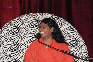 A smiling man with long, swept-back black hair and a small red bindi sits before a zebra-striped cushioned backdrop with deep red velvet drapes behind it. He wears a flowing saffron-orange robe and speaks into a microphone on a stand that curves across the lower frame. Swamiji (probable): long swept-back hair, saffron-orange clothing and a satsang-style setting with a microphone, consistent with an appearance-based label for the page's Swamiji candidate.