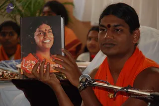 A man in an orange sleeveless garment with a small gold pendant necklace and a red bindi on his forehead holds up a hardcover book titled Living Enlightenment, which shows a smiling woman in orange with a red bindi and beaded necklace. He is seated in front of a microphone on a stand. Behind him, several people in orange clothing sit in a room with white draping and potted plants. Several seated people are visible in the background. A dark tray with colourful objects sits at the left edge.
