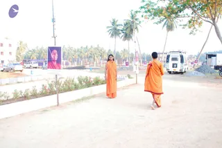 Outdoor photograph of a dirt road lined with a low flower bed of red blooms and palm trees. Two people in saffron-orange robes stand on the road: one facing the camera with long dark hair and a draped orange wrap, the other walking away in an orange robe with maroon trim. A purple-framed poster on a pole shows a smiling woman's portrait. A white van, parked cars, a construction site with gravel and a dark hall stand in the background.