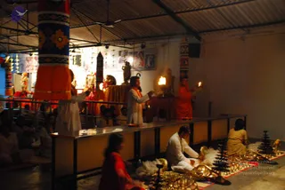 A photograph of an evening puja in a covered hall with a corrugated roof and a metal railing. A standing priest in white robes and an orange stole raises a lit flame over a shrine decorated with orange and red cloth and garlanded deity murtis. To the right, a figure in an orange robe holds a lamp near a lit oil lamp on the altar. Brass oil lamps and a small brass object sit on the floor in front, where a seated man in white clothing tends the ritual items. A blurred woman in a red sari sits in the foreground, and a seated audience in white watches from the left behind a railing. Painted tall pillars with bold geometric colours frame the scene.