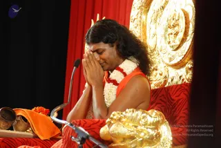 A person with long dark hair sits with palms pressed together at chest level in a namaskar gesture, eyes closed, on an ornate gold-and-red throne. He wears an orange sleeveless garment and a white flower garland with a red-and-white bead necklace. A microphone stand is in front of him, and a brass vessel, an orange cloth and orange flowers rest on a table at left. A large gilded carved backrest with figurative relief stands behind him against a red drape. Visible appearance cues include long swept hair and orange clothing, but the page does not state he is present, so the identity is uncertain.