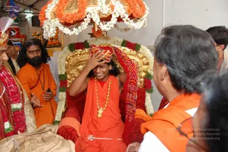 A photograph inside a temple with a gilded throne-like seat draped in red and white flower garlands, under a canopy of orange and white marigold garlands. A smiling man in orange saffron robes and a gold necklace with a pendant sits on the seat, while another person in red and gold cloth presses a red and gold cloth turban onto his head. To the left, a man with long dark hair and a full beard wears an orange robe and a rudraksha-style mala, and a figure in red garlands stands at the edge. Foreground shows a man in an orange shawl and a woman seen from behind, with a brass trident on the floor and a colourful painted wall in the background.