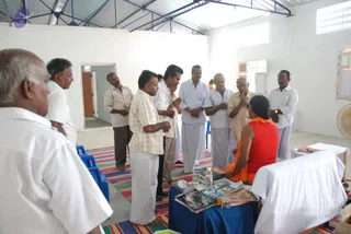 Inside a plain white-walled hall with a corrugated roof and a striped mat on the floor, a group of men in white dhotis and shirts stands in a semicircle with hands folded in greeting. Facing them is a seated figure in an orange-saffron robe with dark hair, seen from behind, at a blue-draped table holding books, magazines and water bottles. A man in a checked shirt and dhoti stands at the left, and an electric fan sits at the right edge. The scene reads as a reception or darshan-style greeting at a small gathering space.