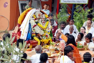 Outdoor temple-style ritual in which a deity murti is decorated with white and yellow marigold garlands and a white cloth, seated on a red and gold palanquin. In the foreground, a person in an orange robe with a bare arm raised stands before an offering table laden with bananas, a carved watermelon, brass vessels, a kalasha and fruit baskets. Around them, devotees in white clothes and yellow lanyards press hands together in anjali, while a woman in a red blouse and a man in a white shirt watch nearby. Nearby hangs a banner with a timing notice and a framed portrait, with orange-painted walls and green shrubs behind.
