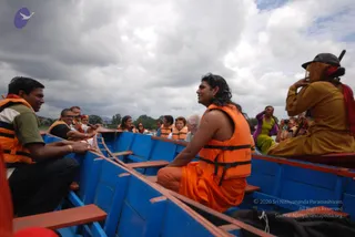 A boat ride on a lake under a cloudy sky. Blue wooden boat with red trim carries a group of passengers wearing orange life vests. In the centre, a person with long dark hair wears an orange life vest over orange robes and sits on the side bench, smiling and looking to the left. A bearded older man in a white shirt sits nearby. On the right, a person in mustard-yellow robes and a dark cap holds a camera, and a woman in a lime-green top raises a hand. A man in a dark shirt and orange life vest sits at the left edge. Distant trees and hills appear across the water.