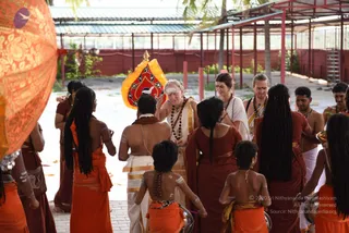 A courtyard ritual scene under a corrugated roof with red pillars and a red-walled compound behind. A man with white-painted forehead and a white-and-gold dhoti, wearing layered rudraksha-style beads, stands centrally with his hand raised toward a devotee. Nearby, a woman in a white top and a man in a white dhoti with beads stand among others, while several people in orange and maroon saffron-toned garments, some with long braided hair, gather around. A large orange and gold ceremonial umbrella fills the left foreground, and a red-and-gold ceremonial banner with a white bird motif hangs behind. Some attendants hold metal bowls. Several bare-backed young participants with white body markings stand in the foreground.