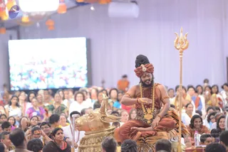 A seated man in saffron-orange robes and a matching turban with a jewelled ornament sits on a tiger-skin-covered seat beside a gold bull statue (Nandi). He wears long gold necklaces, a rudraksha-style gold pendant, bangles and a beard, and holds his hands together in front of him while addressing a large seated audience. A golden trident stands to his right. Behind him a large screen shows a crowd, and a few people in white and colourful saris sit in the foreground audience. Warm lanterns hang from the ceiling. Swamiji (probable): the seated speaker's saffron robes, turban, long gold necklaces and tiger-skin seat match the page's Swamiji reference, so the speaker is identified as a probable in-person Swamiji giving a discourse.