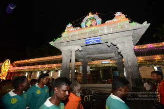 Night photograph of a temple gateway at the Arunachaleshwara Temple in Tiruvannamalai, a grey stone gopuram-style entrance decorated with colourful fairy lights and a painted panel of deities above. A blue sign on the pillar reads Arunachaleshwara Temple. Several young men in teal-green polo shirts with yellow logos stand in the foreground, and a bearded man in an orange-saffron robe walks among them. Further buildings are strung with pink and multicolour lights, and a lit figure stands at the left edge.