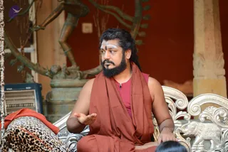 A photograph of a bearded man seated on an ornate silver-finished throne-style chair, speaking and gesturing with his right hand open. He wears a rust-red unstitched wrap over a magenta sleeveless top, with white and red tilak markings on his forehead and a long dark braid. Behind him stands a bronze dancing Shiva-style figure against a red wall, and a leopard-print cloth lies on the chair beside him. A person's head is partly visible at the lower right, and a small microphone clip is visible on his chest. Swamiji (probable) appears in the centre, based on the saffron-red sannyasi-style wrap, long hair and forehead markings on a ceremonial seat; he is shown as an in-person figure at a courtyard-style session.
