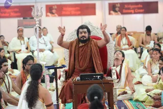 A bearded man in a rust-orange robe and orange shawl, with a long rudraksha mala and gold necklace, sits on an ornate silver-and-red throne, arms raised mid-discourse. A silver trident stands at left and a microphone sits on a wooden lectern. Around him, seated participants wear white dhotis and shawls, some with orange sashes and rudraksha malas; a man in an orange-and-gold shawl sits beside the throne with hands in a meditative pose. Banners behind read Nithyananda Padukas Mantra, Nithyananda Padukas Shiva Yoga, and Nithyananda Satsang Center. The setting appears to be a large hall used for a satsang or initiation session. a person: the seated speaker wears saffron-orange robes, rudraksha mala and ceremonial gold necklace on a throne.
