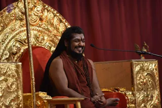 A seated man with long dark hair and a full beard smiles broadly on an ornate gilded throne with red velvet cushions, in front of a deep red curtain. He wears a rust-red sleeveless wrap over bare shoulders, with rudraksha malas hanging across his chest and a red-and-gold necklace. A vertical tilak marks his forehead. He sits cross-legged with hands folded, and a microphone stand stands to his right beside a small gilded carved stand. One physical person is visible.