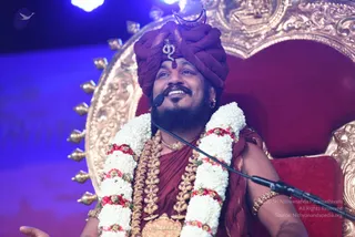 A man seated on an ornate gold-and-red throne with a carved golden backrest, speaking into a microphone on a stand. He wears a deep maroon turban with a silver ornament at the front and a crown-like silver headpiece, a maroon shawl draped over a maroon garment, and multiple layers of gold necklaces. Thick white and red flower garlands hang across his shoulders and chest, and he has gold bangles on his wrist. His right hand is raised open in an expressive gesture while he smiles and looks upward. Blue stage lighting glows behind him. The turban, throne and maroon attire with ornate gold ornaments and garlands match prominent a person cues, so the appearance-based label is probable.