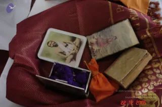 A close photograph of ritual objects laid on a maroon silk cloth with gold border. A white square tin lid shows a printed image of a bearded seated figure in white clothing. Beside it lies a small flat book with a faded pale cover. In a silver rectangular box filled with purple flower petals or fabric pieces. An orange drawstring pouch lies on the cloth, and a worn, tan-covered book rests at the right edge. No people are visible.