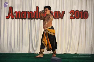 A performer stands on a green stage mat in front of a white draped curtain with large red lettering reading Anandasav 2010. He is bare-chested and wears a black and gold-yellow dhoti-style lower garment with a gold waist belt, ankle bells, and a long rudraksha-style bead necklace. His face is painted with a red bindi and his short hair is styled upward. One arm is raised to the head in a classical dance pose while the other hand rests at the waist, with a gold armlet on the upper arm.