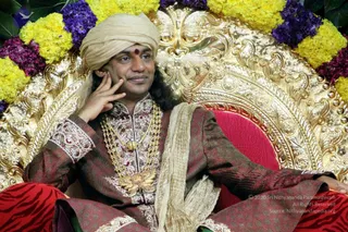 A seated man sits on an ornate golden throne with a red cushion, framed by garlands of yellow marigolds and purple flowers. He wears a beige turban, a small red bindi on his forehead, a maroon and gold embroidered sherwani, layered gold necklaces and a beige shawl draped over one shoulder. He rests one hand against his cheek and looks off to the side with a slight smile. Red patterned fabric covers his legs. The scene appears to be a formal ceremonial setting.