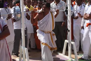A man in white dhoti and white shawl with gold-yellow border lifts a barbell across his shoulders in an outdoor setting with a white squat rack. He has a red tilak mark on his forehead and a rudraksha-style mala visible at his neck. Behind him a crowd of men in white shirts and white dhotis, some in saffron-orange garments, watch and clap. A man in orange cloth stands among the spectators. Bare feet stand on a sandy ground with a purple mat beneath the rack.