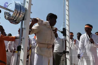 Outdoor photo under a clear blue sky of a man in a cream kurta and dhoti with a tan leather belt lifting a heavy barbell on a white steel rack with chrome weight plates. Behind him, a crowd of men in white dhotis and kurtas and one in orange robes watch. On the right, a man in white kurta with a orange headband and rudraksha mala holds a microphone and gestures. Another man in an orange headband stands nearby. Text overlay in the lower right reads copyright and source notice, which is not included.
