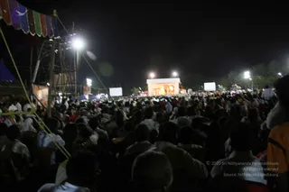 A large nighttime outdoor crowd fills the foreground, with many heads and shoulders of people seated or standing and facing a brightly lit stage at the far end of a open ground. The stage is decorated with warm orange and gold lights and a red-and-white canopy, with two white projection screens flanking it. Tall floodlights on poles illuminate the scene, and a striped tent canopy in red, green and blue hangs over the left edge. Yellow guy ropes run through the frame. The crowd is dense and appears to number in the thousands, though only individual heads are clearly distinguishable up close. No individual figure on the stage can be clearly made out.