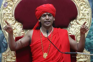 A man seated on an ornate gold-carved throne with a deep red velvet back speaks into a microphone while raising both forearms with index fingers pointed upward. He wears a red turban with a small red bindi, a red saffron-style shawl draped over bare shoulders, a string of white beaded necklace, a gold pendant, a long gold bead mala, and a beaded bracelet. The setting is a stage with a green backdrop.
