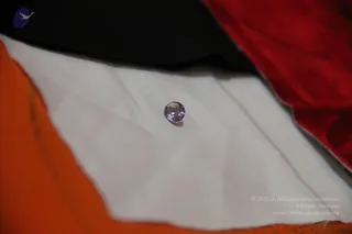 Close-up photograph of a single small round faceted lavender-purple gemstone resting on a folded white cloth. The white cloth sits between a dark black fabric at the top and an orange fabric at the lower left, with a red velvet-like fabric at the upper right. No people are visible.