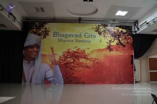 A photograph of an empty stage hall with a large printed backdrop banner reading Bhagavad Gita Dhyana Satsang. The banner shows a smiling man in a pale blue turban and pale blue long kurta with a gold-toned ornament, raising one hand in a greeting, beside a red and orange autumnal tree scene and a faint carved temple relief. The banner appears to be the only figure in the room; no physical people are visible in the hall. Black curtains frame the stage, and a white glossy floor reflects the backdrop.