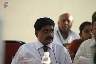 Indoor seated audience at a meeting or satsang. In the foreground a man with short black hair, a moustache and a red bindi wears a white collared shirt, striped tie and lanyard, looking to the side. Behind him an older bald man in a white shirt and lanyard sits, and a woman in an orange sari with gold earrings watches. A water bottle and glasses sit on the table in front. A pale yellow wall forms the backdrop. No one present is shown in a leader's role or in distinctive a person attire.