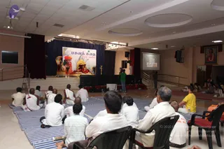 A seated audience of mostly men faces a stage in a large hall with striped blue-and-white mats on the floor and plastic chairs at the front. Many are dressed in white or cream clothing. On the stage backdrop is a banner reading a Tamil title, with a large portrait of a figure in orange robes and a crown-like headpiece, flanked by golden ornaments and a golden throne-like seat with flowers. A cameraman in green stands at a tripod filming the stage, and a projector screen on the right shows a slide. Ceiling fans and recessed lights are visible overhead.