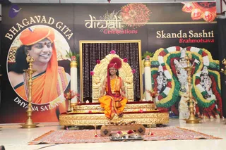 A photograph of a stage set for a Bhagavad Gita satsang. A seated figure wearing an orange dhoti and a deep red turban, with a red shawl and a gold chain, sits cross-legged on an ornate gold throne-like seat with a red cushion, facing forward with a small oil lamp and a brass tray of flowers before him. A large framed backdrop shows a smiling man in an orange turban and orange shawl with a rudraksha mala, labelled Bhagavad Gita and Dhyana Satsang, beside a banner with fireworks reading Diwali Celebrations, and a panel of Skanda Sashti Brahmotsava with a decorated deity image garlanded in red and white flowers. Tall brass lamps stand on either side and a patterned rug covers the floor.