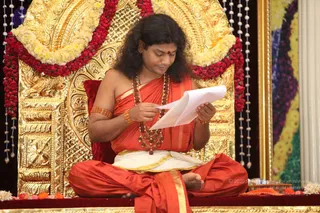A seated man in saffron and orange robes sits cross-legged on an ornate gold throne with a red cushion, decorated with red and yellow flower garlands and bead strings. He wears a red bindi, rudraksha-style mala necklaces, gold bangles and a gold-bordered white dhoti, and reads from a sheet of white paper held in both hands. Fresh flowers and fruit rest on the red platform in front of him. The setting is a temple-style hall with gold and floral decor.