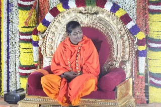 A single person sits cross-legged on an ornate gilded throne with a deep red cushioned seat and red bolster pillows. The seated figure wears an orange-saffron shawl draped over the shoulders and orange cloth on the lower body, with a beaded rudraksha-style mala necklace and a red dot on the forehead. The throne stands in front of a backdrop decorated with garlands of white, yellow, red, purple and green flowers. A black audio speaker sits at the lower left. Appearance cues are a shawl, saffron clothing and a rudraksha mala, so this is a person candidate seated in the presence of a throne display.