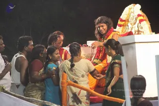 Night photograph of an outdoor darshan at a raised platform. A seated figure with long swept-back dark hair, wearing an orange-saffron garment and a red and white garland of flowers, sits on a red-cushioned seat before a golden ornate throne decorated with white and red flowers, holding a small white bowl and reaching toward a devotee. Several devotees in saris and casual clothes stand before the platform: a woman in a cream floral sari, a woman in a dark green sari, a girl in a blue dress, and men in white and red tops, with a man in a red garment. An orange barrier rope runs along the front. The seated figure has distinctive long hair, orange clothing and a flower garland, consistent with a probable Swamiji presence; the figure is the only candidate in view.