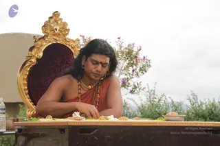 A photograph of a man seated at an outdoor table in front of an ornate gold and crimson velvet throne-style chair. He has long dark hair, a red bindi, gold earrings and a gold pendant necklace over rudraksha malas, and wears an orange-red shawl over his bare shoulder. He is eating from a green leaf plate arranged with fried snacks, a white rice-like dish and small bowls; his right hand is reaching toward the food. A water bottle is on the left and trees with pink flowers and an overcast sky fill the background. Given the page's a person flag and the throne, the appearance cues of long hair, orange-red shawl and malas suggest a person candidate present in person, though identity is uncertain.
